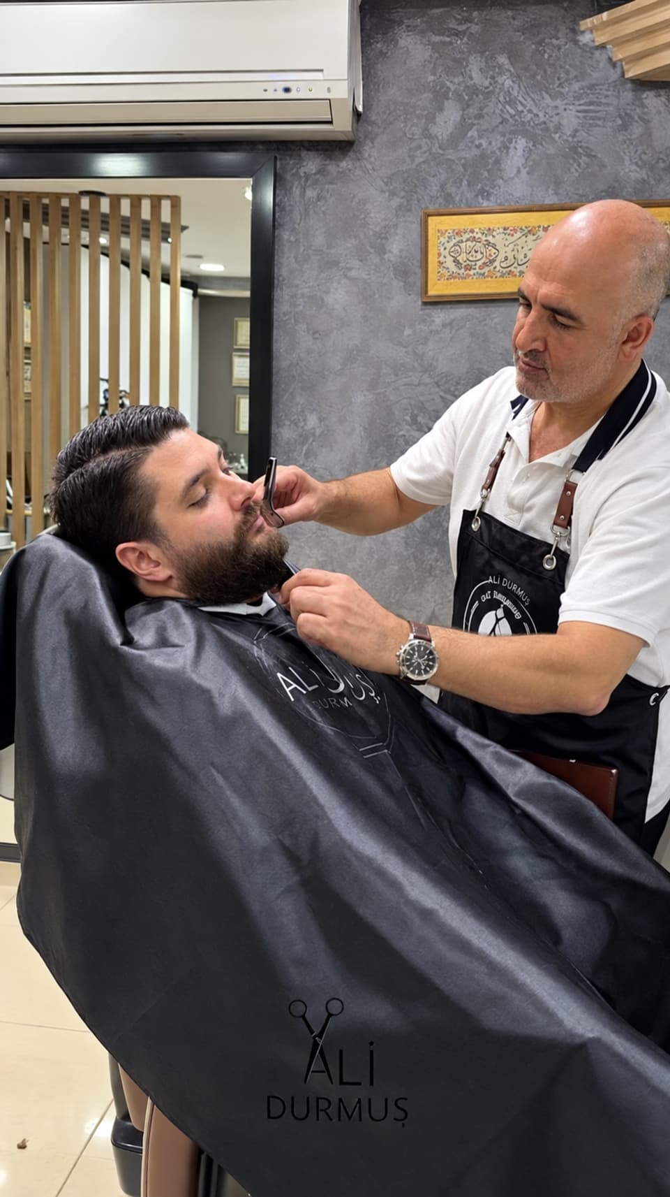 Ali Bey, arkaya yaslanmış bir müşterinin bıyık ve sakalını makasla düzeltiyor